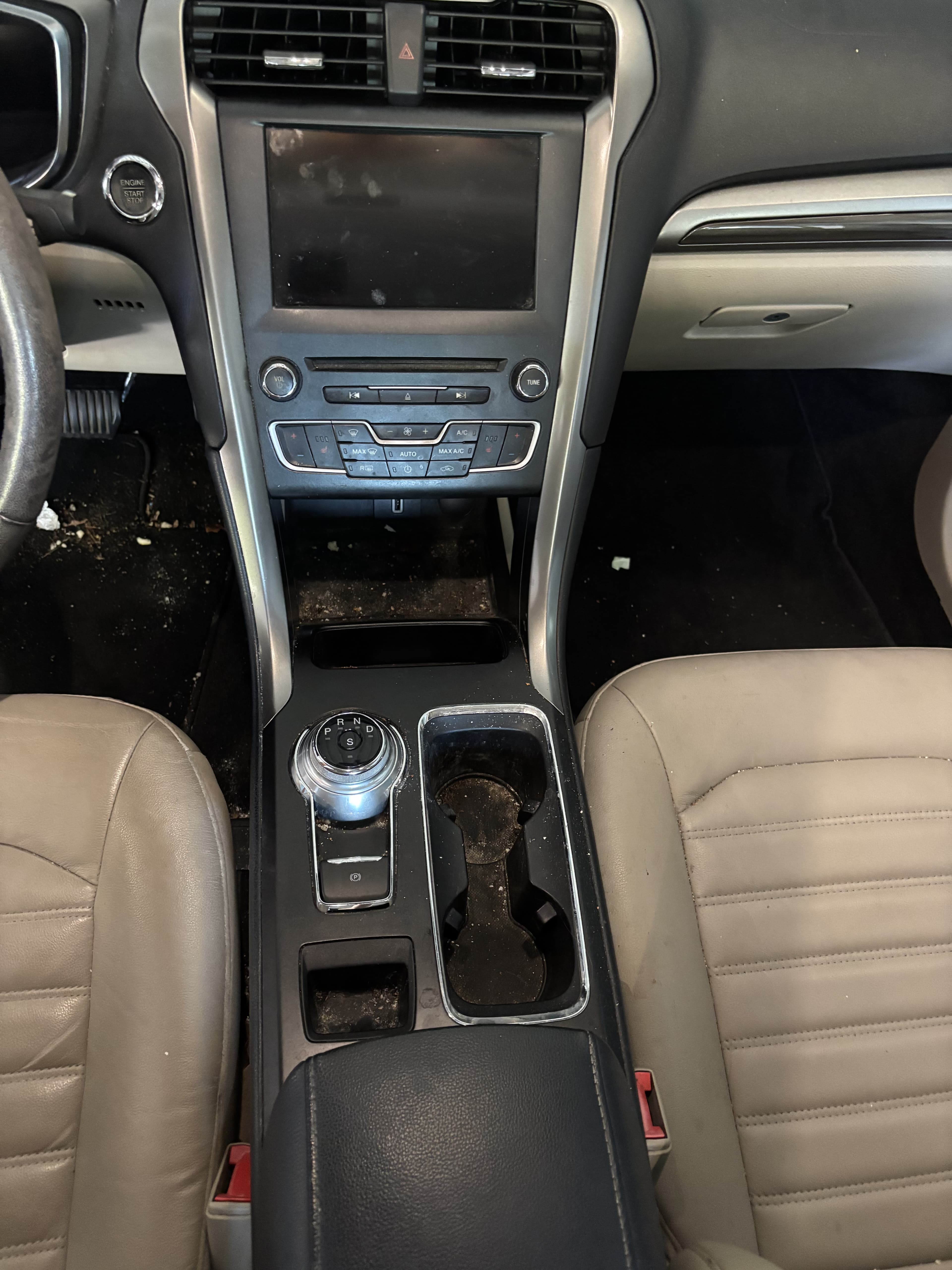 Dirty car interior showing tan leather seats, center console, rotary gear shifter, and floor debris.