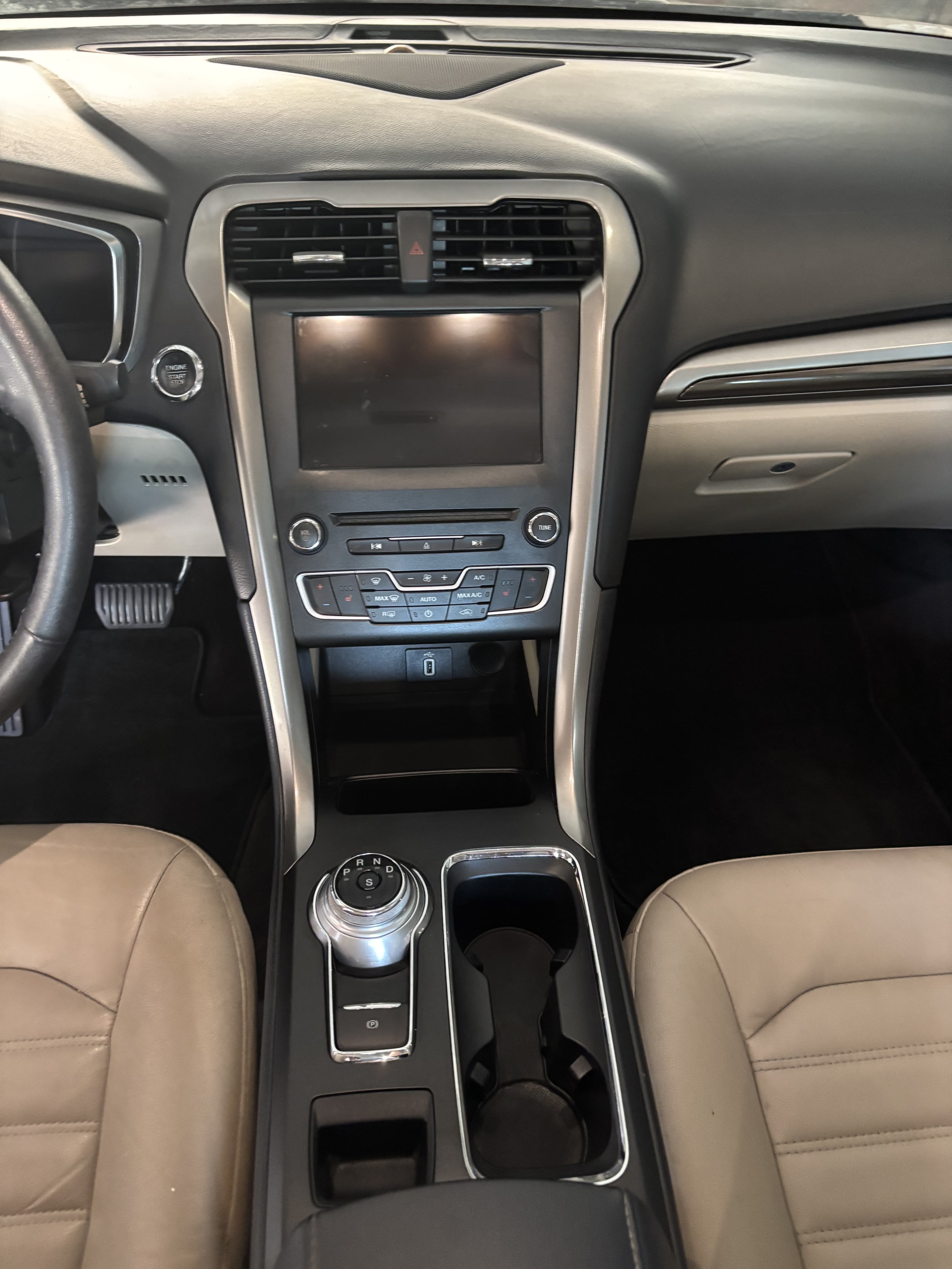 Modern car interior featuring a touchscreen infotainment system, rotary gear shifter, and tan leather seats.